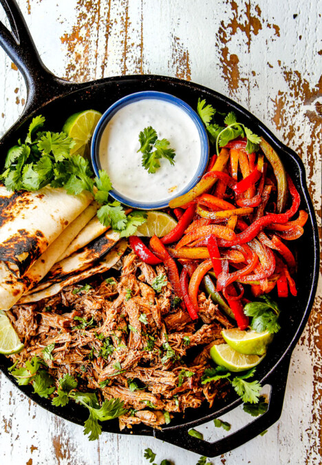 top view of pork fajitas in a skillet with salsa verde pork, bell peppers, onions, limes, cilantro and tortillas