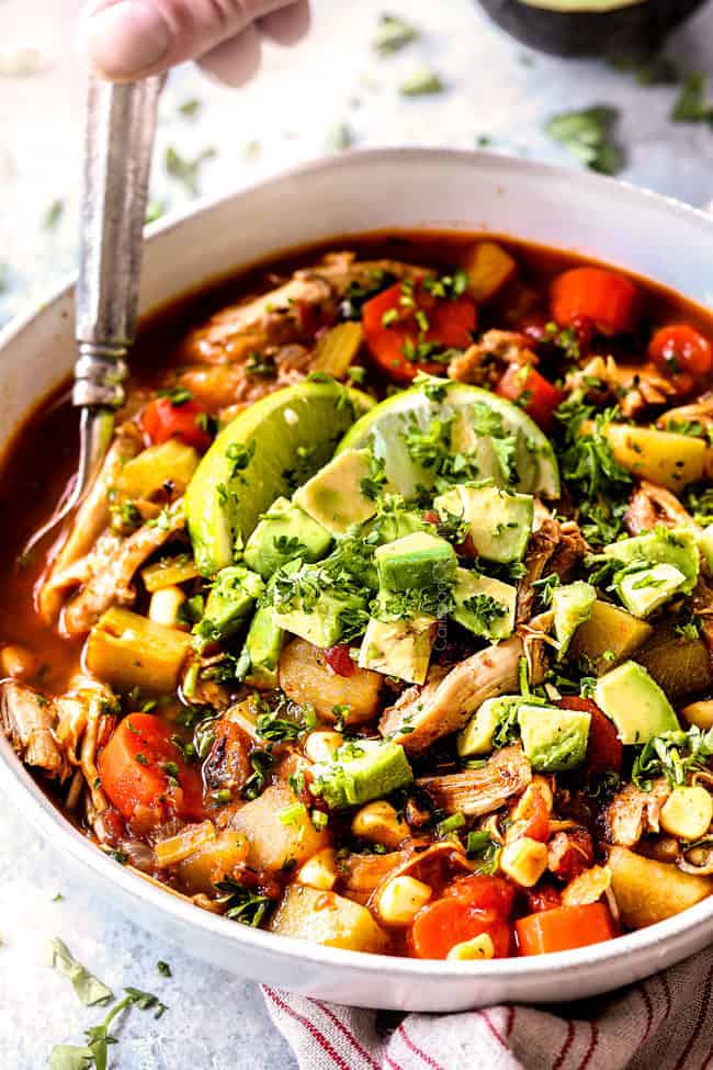 a hand holding a spoon in Caldo de Pollo (Mexican Chicken Soup) with poatoes, carrots, celery topped with avocados
