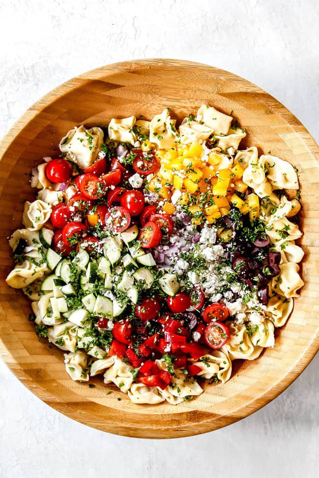 showing how to make Greek Past Salad by adding pasta, tomatoes, olives, bell peppers, red onions, cucumbers to a wood bowl