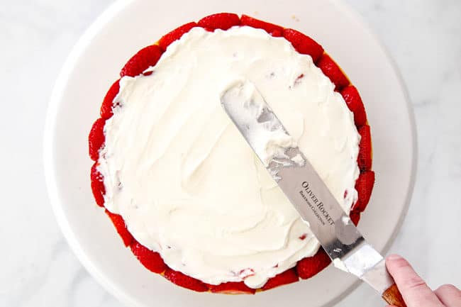 showing how to layer strawberry shortcake cake by adding whipped cream on top of the strawberries