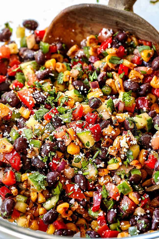 showing how to make black bean salsa by tossing black beans, corn, red bell peppers, cucumbers, jalapenos, red onions, cilantro, lime juice and Cotija cheese together in a glass bowl