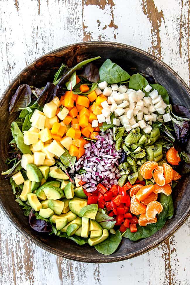 showing how to make shrimp avocado salad recipe by adding avocados, pineapple, mangos, jicamo, kiwis, red onion, bell peppers ad Mandarin oranges to a wood bowl