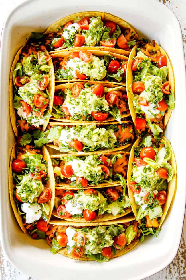 top view of showing how to make baked ground beef tacos by topping with lettuce, tomatoes, guacamole and sour cream in a 9x13 baking dish
