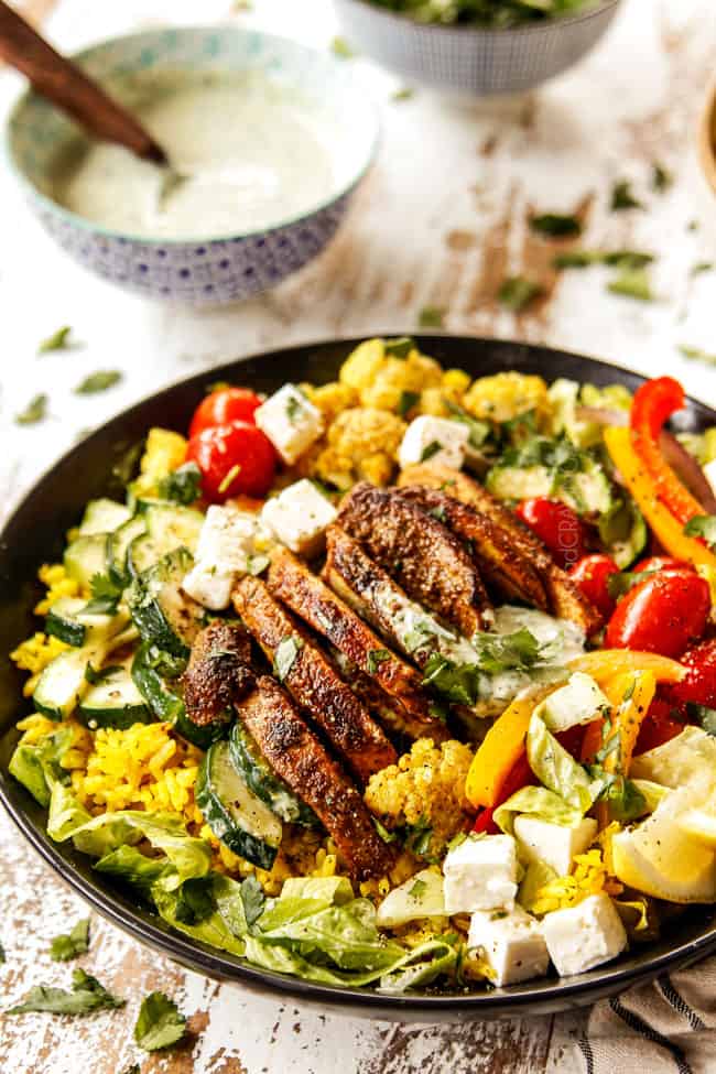 showing to make chicken shawarma into bowls by adding chicken, roasted cauliflower, bell peppers, tomatoes and zucchini to a black bowl with rice and lettuce