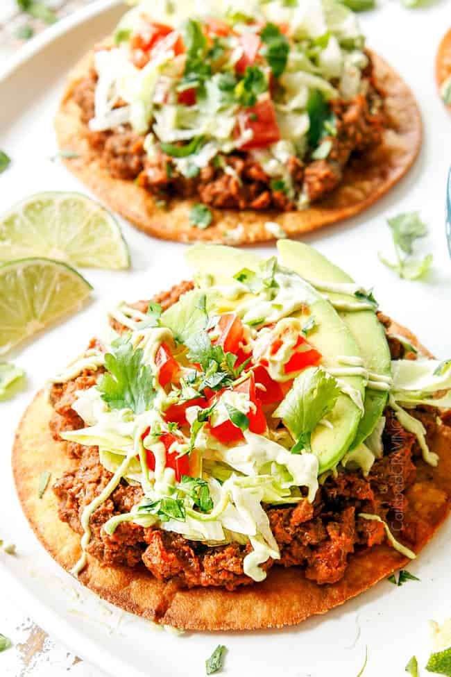 showing how to make best tostada recipe by topping tostada shell with refried beans, beef, lettuce, tomatoes, avocados and sour cream