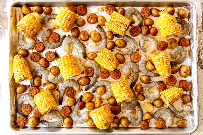 showing how to make sheet pan shrimp boil recipe by adding shrimp to baking sheet with corn, potatoes and sausage and spreading into an even layer