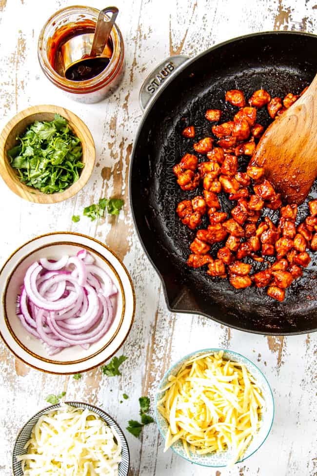 showing how to make BBQ Chicken Pizza recipe by spreading out toppings: BBQ chicken, BBQ sauce, thinly sliced red onions, cilantro mozzarella and Gouda cheese in bowls on a white counter
