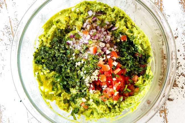 showing how to make guacamole recipe by adding diced red onions, jalapenos, cilantro, tomatoes, salt and lime juice to mashed avocados in a glass bowl
