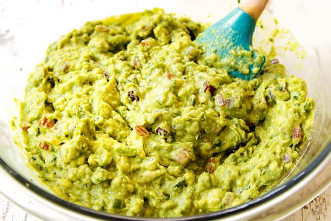 showing how to make guacamole by stirring mashed avocados together with tomatoes, onions, cilantro, lime juice and jalapenos in a glass bowl