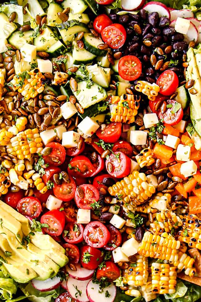 showing how to make Mexican Salad recipe by adding romaine lettuce, corn, black beans, tomatoes, cucumbers, radishes, red onions, tomatoes, cheese, and pepitas to a large wooden bowl
