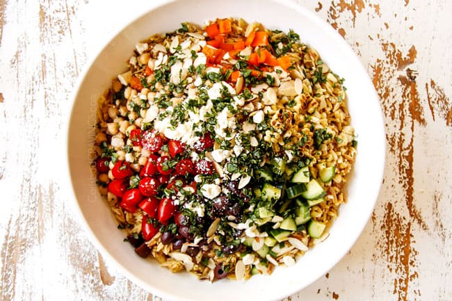 showing how to make orzo pasta salad recipe by adding orzo, cucumbers, bell peppers, tomatoes, olives, chickpeas, basil and feta to a large glass bowl
