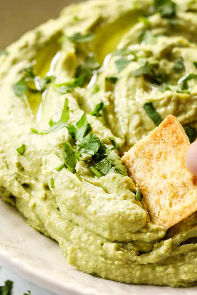 showing how hummus recipe variations by dipping a cracker into avocado hummus in a white bowl