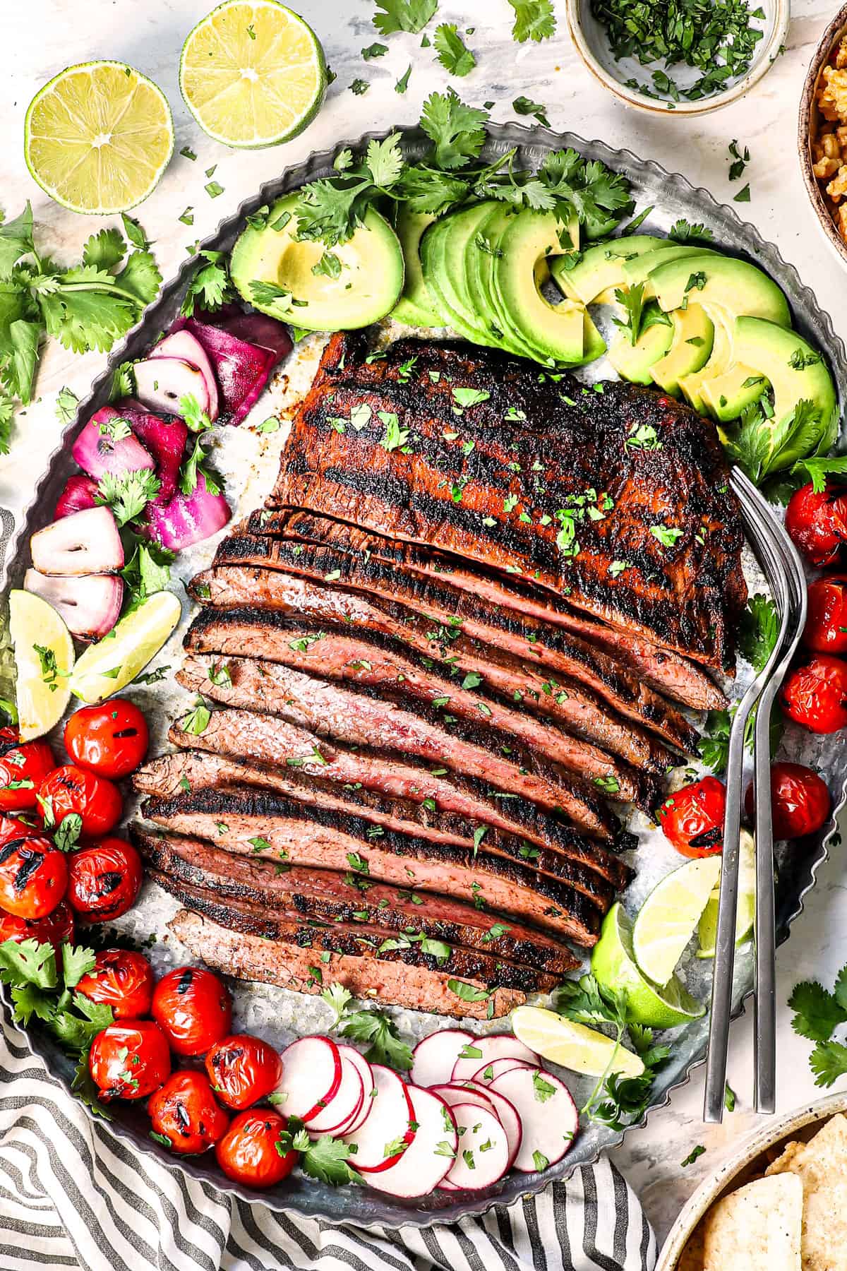 showing how to serve carne asada on a platter with avocados, tomatoes, cilantro, limes, and radishes