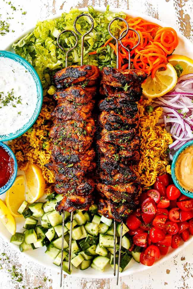top view showing how to serve doner kebab meat by adding kebabs to a plate with hummus, thinly sliced lettuce, thinly sliced red onions, chopped tomatoes, chopped cucumber, curried rice, Lebanese flatbread, chili sauce and garlic yogurt sauce