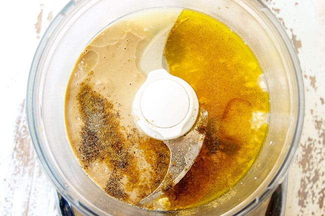 showing how to make homemade hummus by adding tahini, lemon juice, garlic cloves, olive oil, ground cumin and salt to a food processor