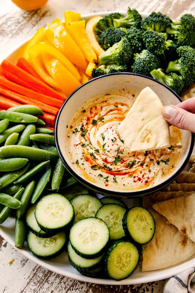 showing how to serve hummus recipe by adding hummus recipe to a black bowl with a platter of broccoli, cucumbers, snap peas, carrots, pita bread and bell peppers