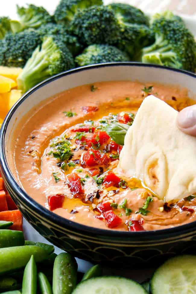 side view showing homemade hummus recipe variation of roasted red pepper hummus in a black bowl dipping a pita into