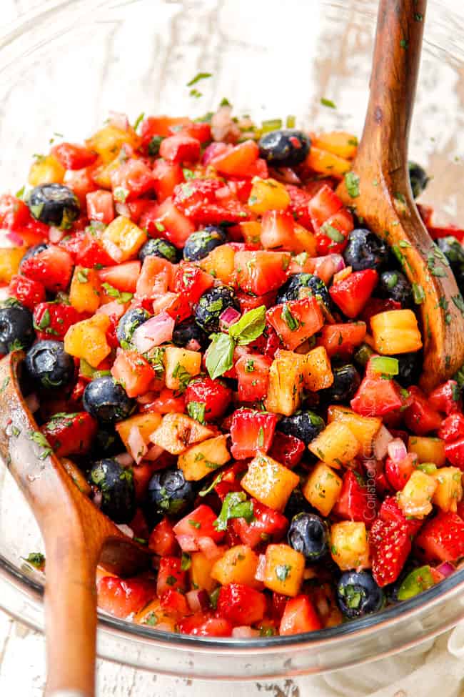 showing how to make strawberry salsa by tossing strawberries, pineapples, cilantro, red onions and cilantro together in glass bowl with two wooden spoons
