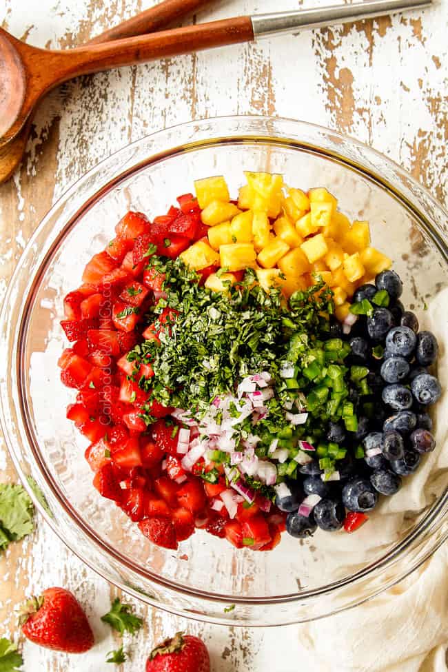 up close showing how to make strawberry salsa by adding strawberries, pineapples, cilantro, red onions and cilantro to a glass bowl