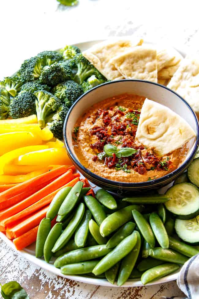 side view showing homemade hummus recipe variation of sun-dried tomato hummus in a black bowl surrounded by snap peas, carrots, yellow bell peppers, broccoli and pita bread wedges