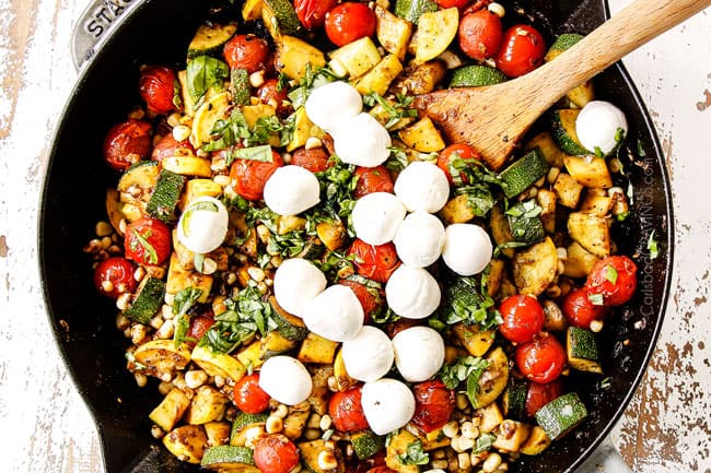 showing how to make Tuscan Chicken by adding fresh basil to the tomatoes, zucchini and corn in a cast iron skillet