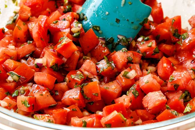 showing how to make Bruschetta Chicken by making bruschetta in a glass bowl with tomatoes, basil, red onions and garlic