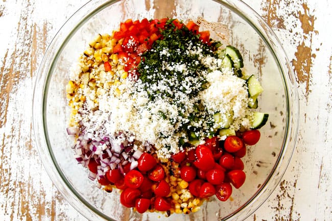 showing how to make Mexican Street Corn Salad (esquites) by combining grilled corn, red bell peppers, tomatoes, cucumbers, red onion, cilantro and Cotija cheese in a glass bowl