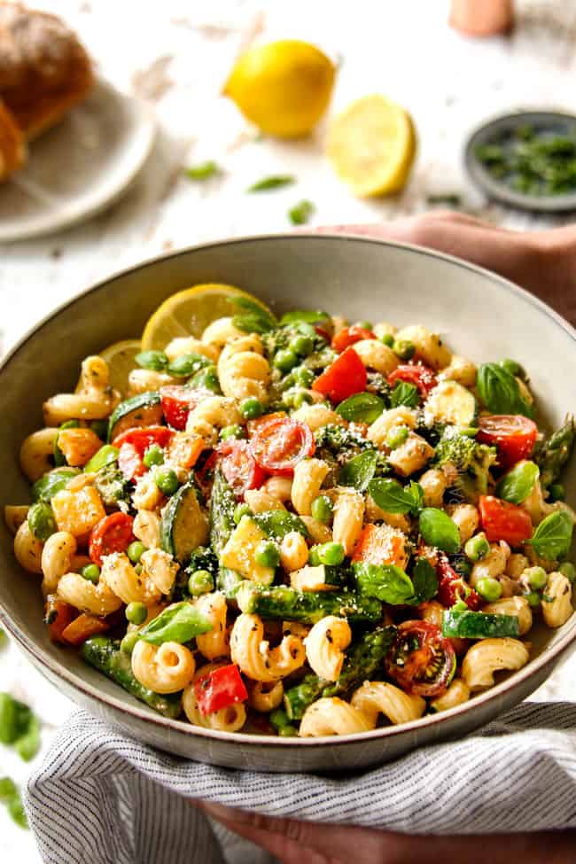 two hands holding pasta primavera recipe in a grey bowl garnished with Parmesan and fresh basil