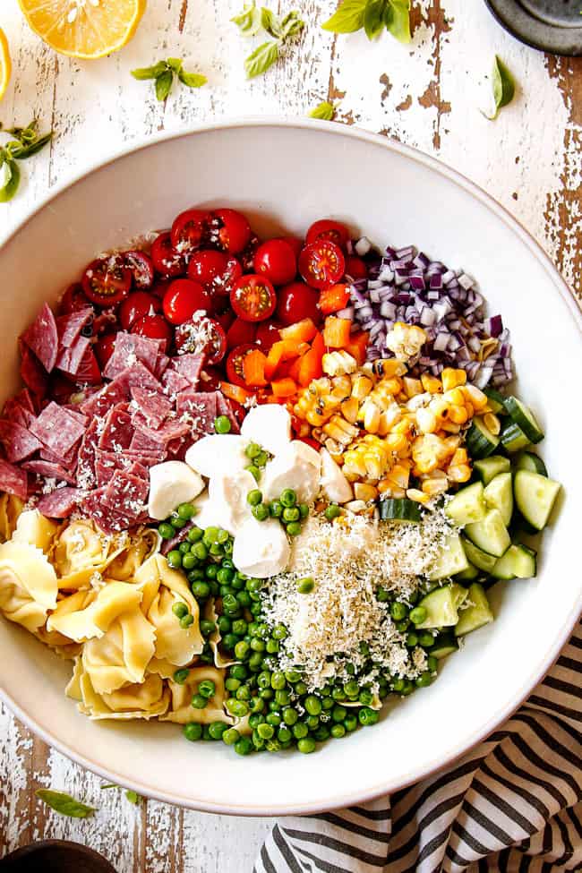 top view showing how to make pesto pasta salad by adding pasta, cherry tomatoes, mozzarella, Parmesan, cucumbers, corn, bell peppers and red onions to white bowl