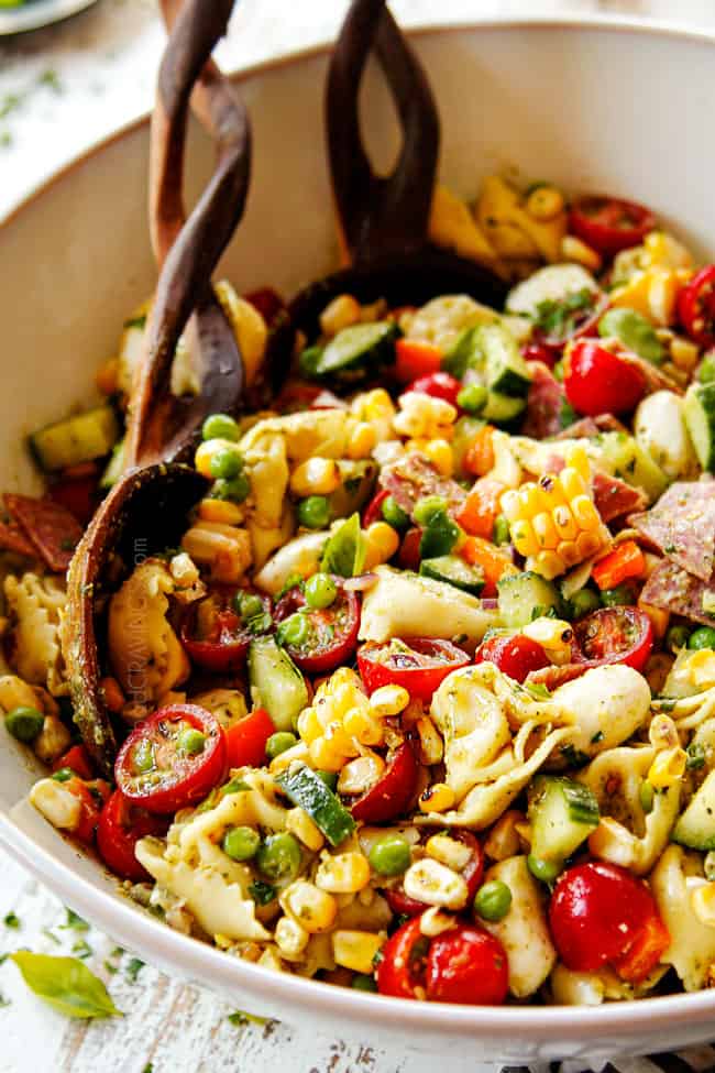 side view of pesto pasta salad recipe with vegetables in a white bowl with wooden tongs