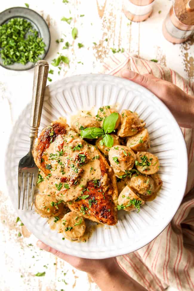 top view of two hands holding a white plate of creamy Tuscan chicken