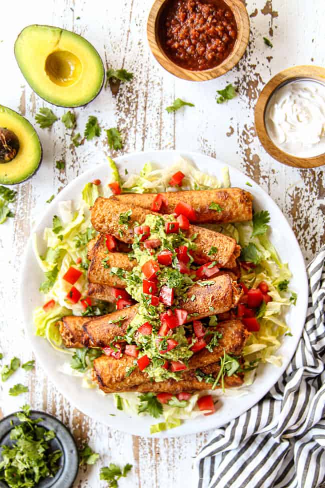 top view of flautas on a white plate with shredded lettuce, guacamole and chopped tomatoes