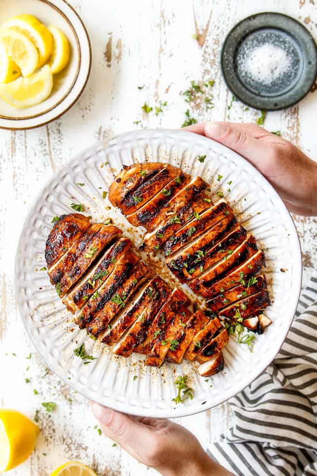 top view of marinated grilled chicken breasts sliced on white plate being held by two hands