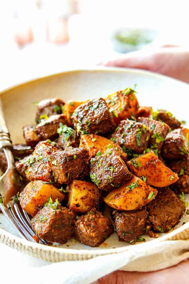 two hands holding steak and potatoes recipe on a tan plate with a silver fork