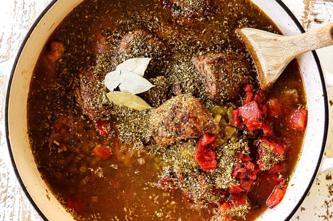 showing how to make pozole by adding chicken broth, diced tomatoed, Mexican oregano, ground cumin and bay leaves to the pork in a white Dutch oven
