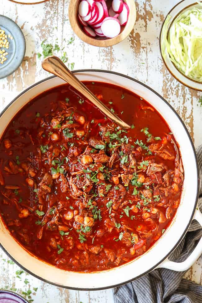 top view of pozole in white soup pot surrounded by bowls of garnishes: cilantro, limes, red onions, radishes