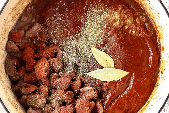 showing how to make Chile Colorado recipe by adding chile sauce, beef, oregano, cumin, bay leaves and beef broth to Dutch oven