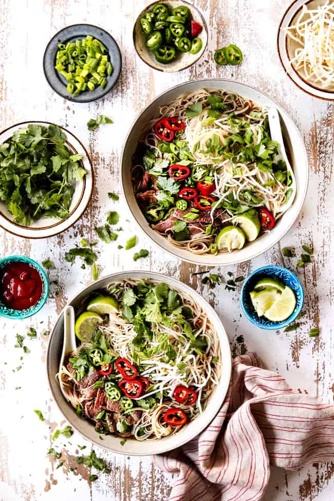 showing how to serve pho with two bowls of soup surrounded by garnishes: cilantro, basil, green onions, bean sprouts, lime wedges, hoisin and srircha