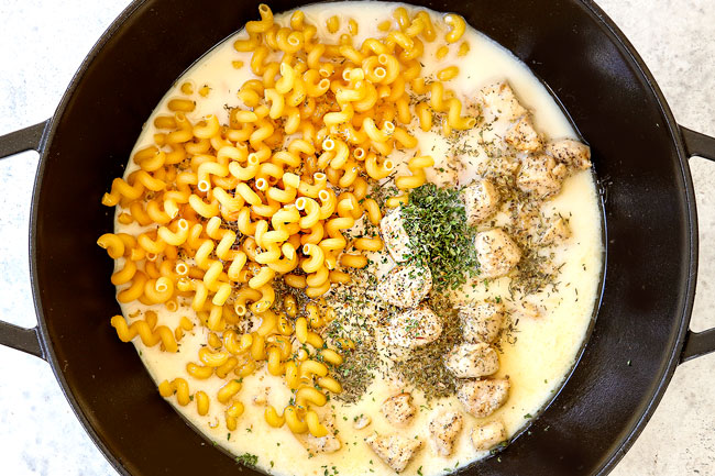 showing how to make chicken pesto pasta recipe by adding pasta, evaporated milk, chicken broth and seasonings to black Dutch oven