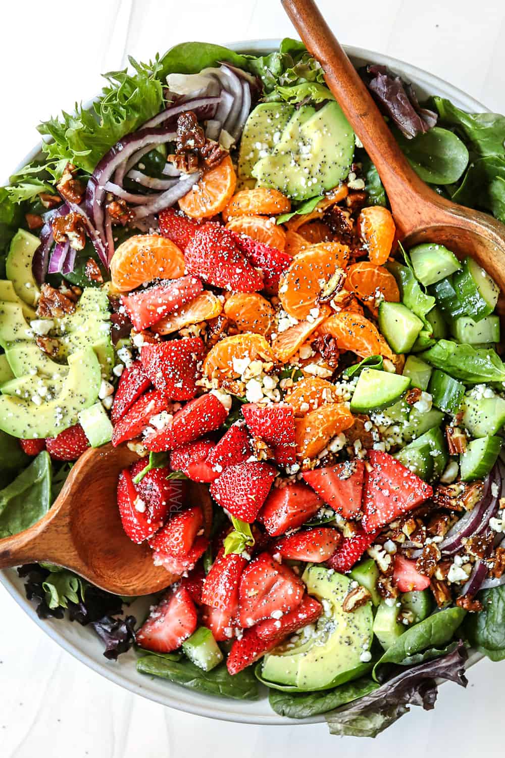 showing how to make strawberry spinach salad recipe by adding spinach, strawberries, clementine oranges, cucumbers, avocados and red onions to a bowl
