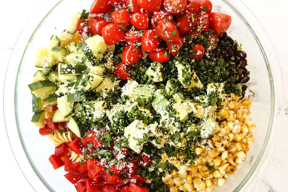 showing how to make Mexican Pasta Salad by adding all of the pasta salad ingredients (tomatoes, cucumbers, bell peppers, black beans, jalapenos, cilantro, corn, Cotija Cheese) to a glass bowl