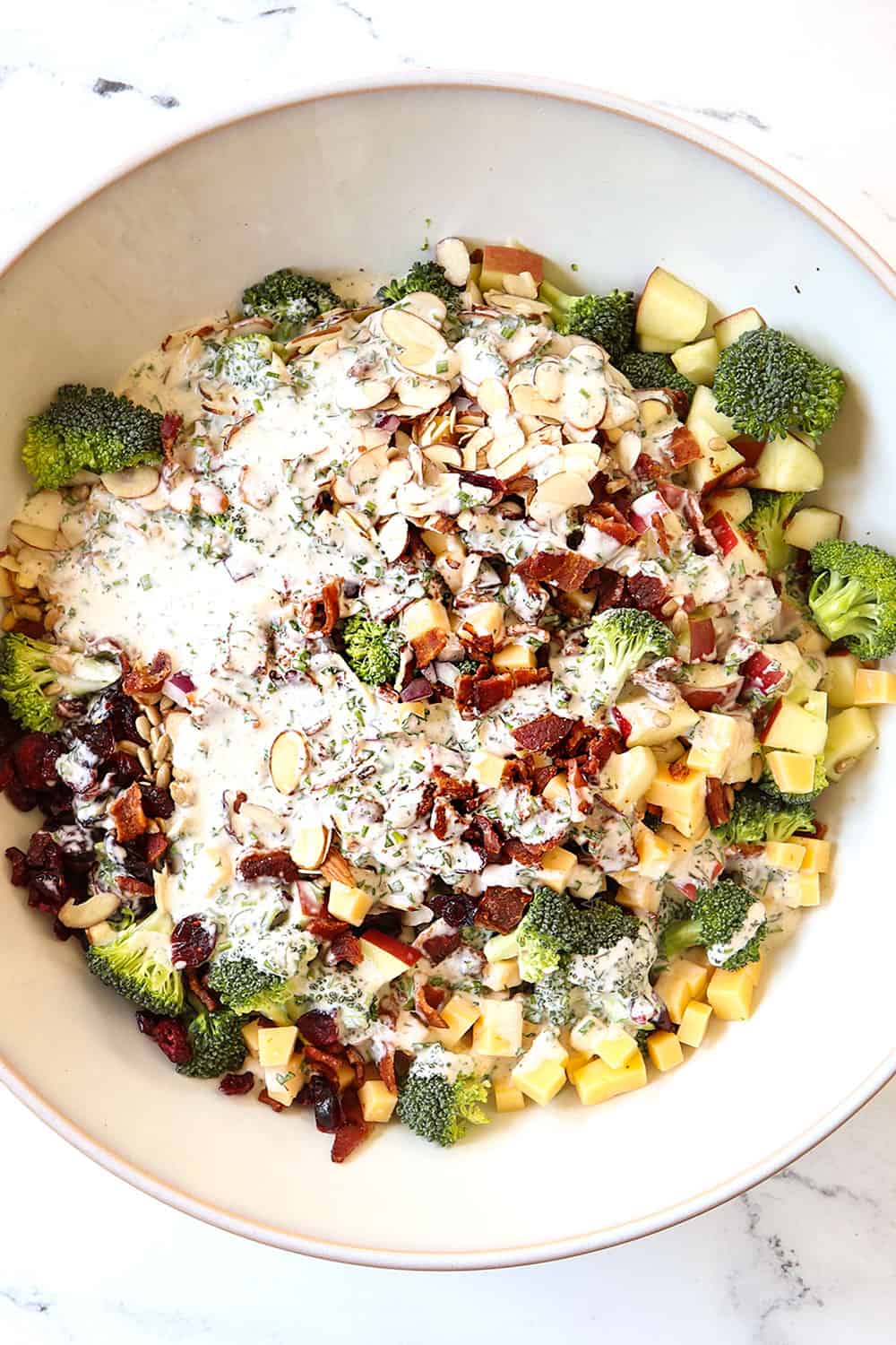 top view showing how to make broccoli salad recipe by drizzling broccoli, bacon cheese, apples with dressing in white bowl