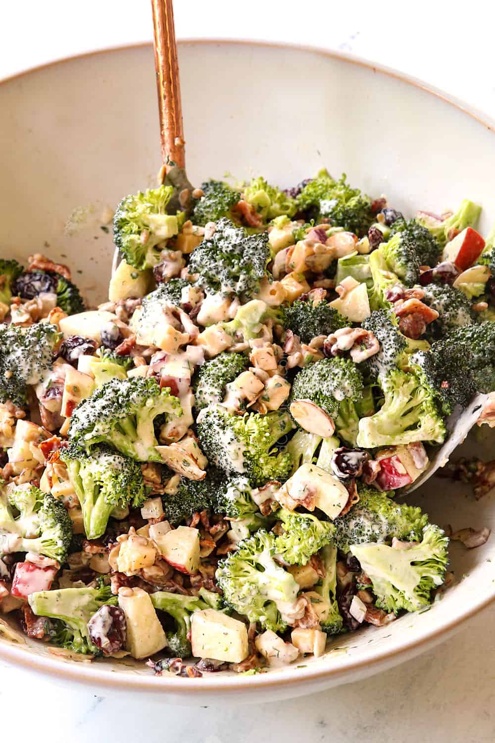 showing how to make best broccoli salad recipe by tossing the broccoli bacon, cheese and apples with the dressing