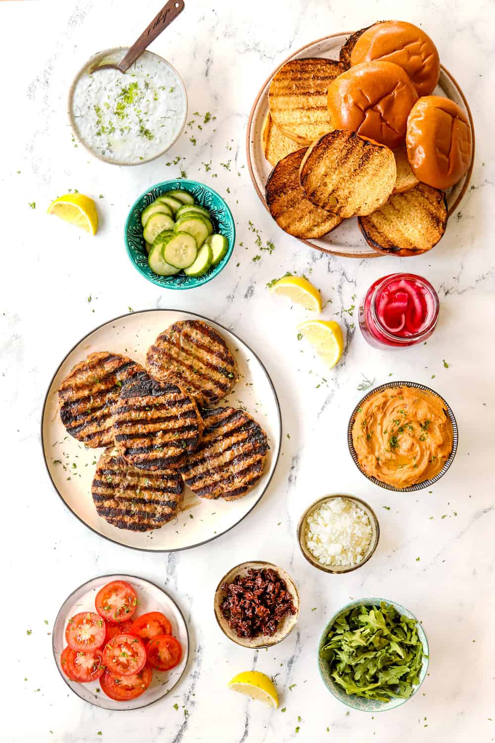 showing how to make Greek lamb burger recipe with top view of lamb burger patties and toppings: hummus, cucumbers, red onions, tomatoes, sun-dried tomatoes, tzatziki