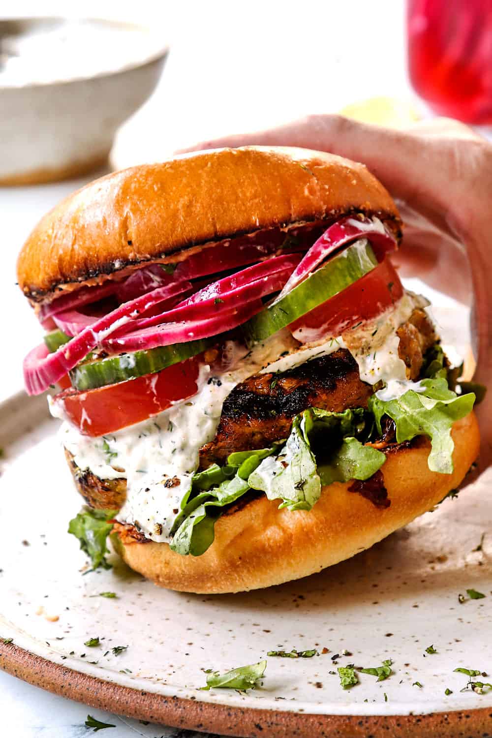 showing toppings for lamb burger Greek style by topping buns with arugula, lamb patties, tomatoes, cucumbers, tzatziki and red onions