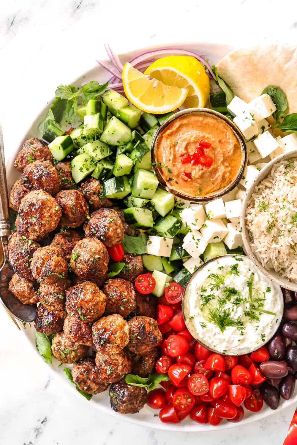 showing how to serve Greek Meatballs (Keftedes) with a top view of a meze platter with cucumbers, tomatoes, olives, feta, pitas, tzatziki and hummus