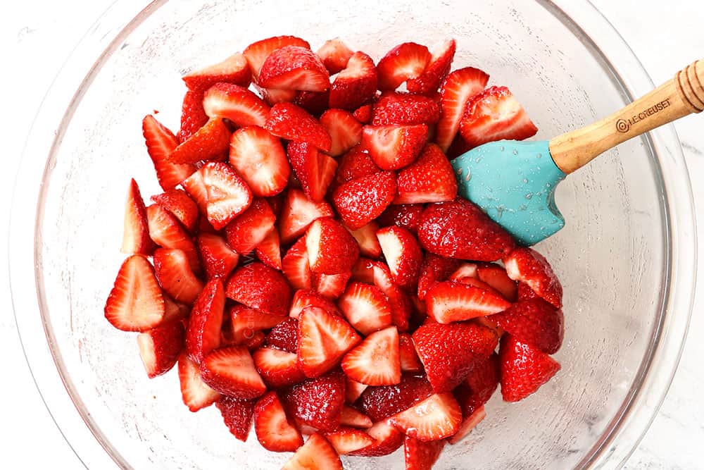 showing how to make strawberry crisp (crumble) by adding the strawberries, lemon juice, sugar and vanilla to a bowl and stirring all together with a spatula