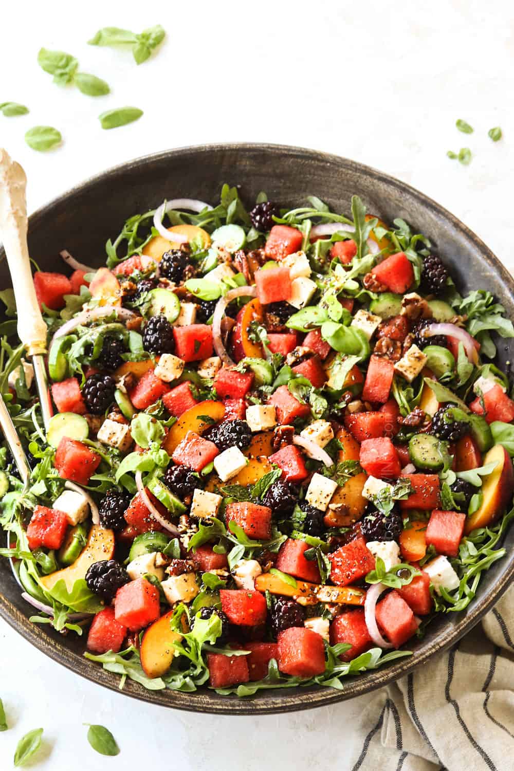 watermelon feta mint salad with basil, cucumbers and arugula in a wood bowl with white and silver serving tongs