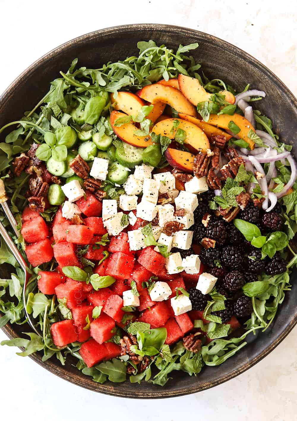 showing how to make watermelon salad recipe by adding watermelon, feta, mint, arugula, cucumbers and berries to a large bowl