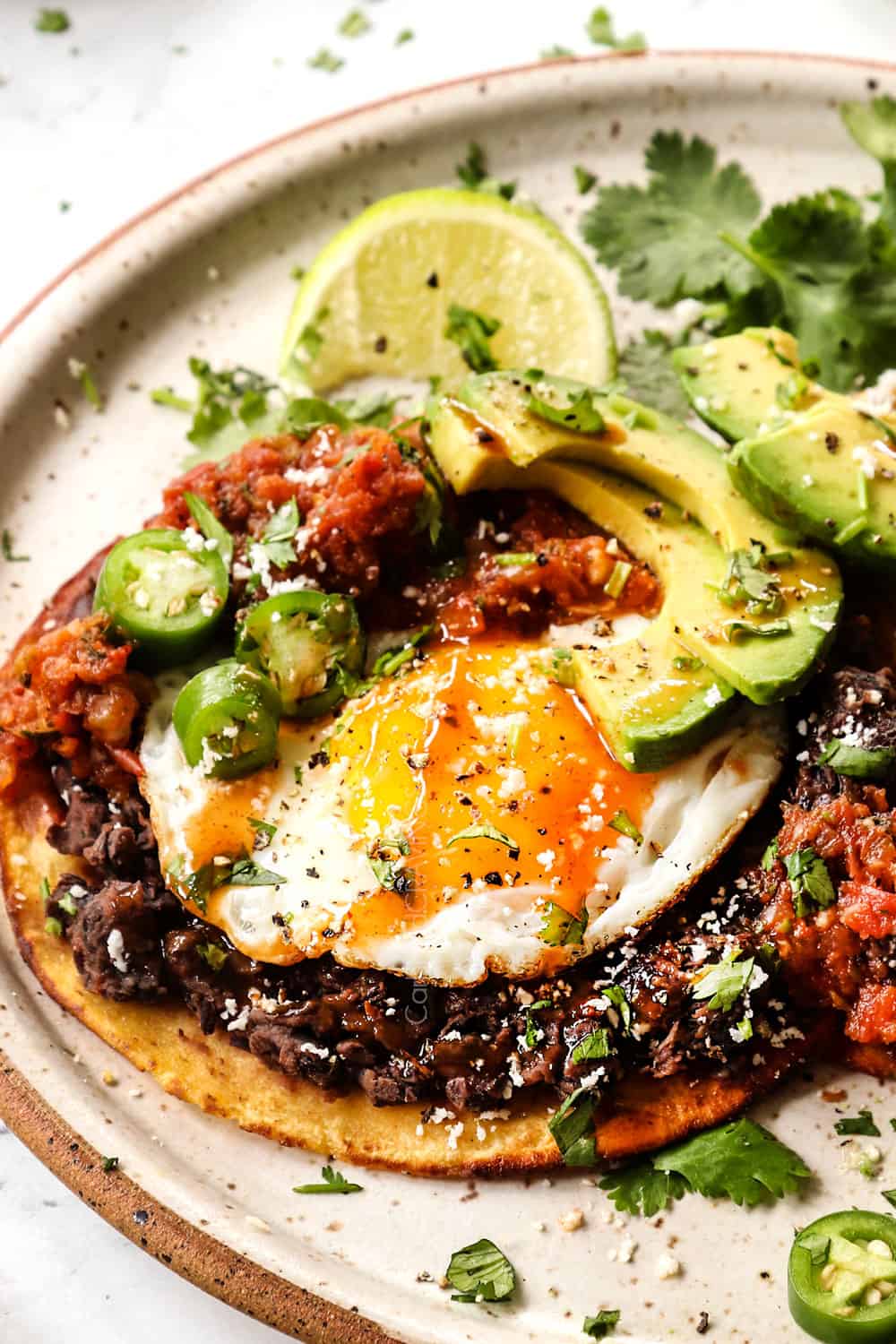 up close of Huevos Ranchers showing how to layer them: corn tortilla, refried beans, fried eggs, salsa and toppings of sliced avocado, Cotija cheese, cilantro and hot sauce